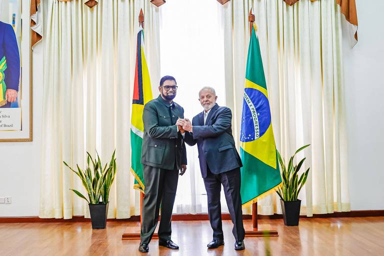 Foto mostra Lula, um homem idoso, branco, de terno, sorrindo e dando as mãos com Irfaan Ali, um homem negro de meia-idade. Atrás deles, as bandeiras dos dois países.