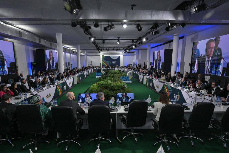 sala de reunião com grande mesa oval e telões na parede, com homens e mulheres reunidos