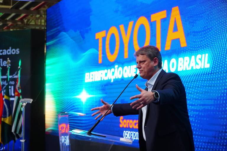Tarcísio está discursando, de terno, em um evento corporativo, com um grande banner da Toyota ao fundo, destacando a presença da marca no Brasil.