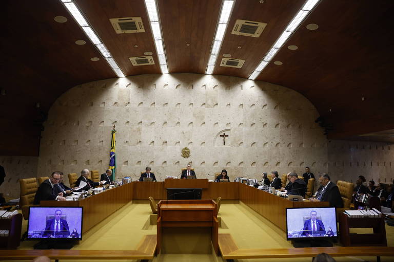 foto mais aberta mostra salão com mesa retangular, ocupada pelos oito ministros e uma tela de cada lado virada para a frente de quem acompanha o julgamento 