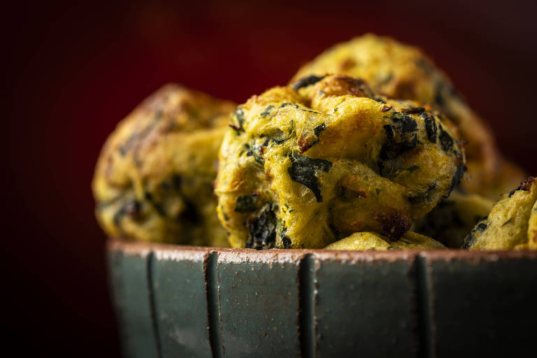 Bolinhos de superfície irregular, na cor amarela com verduras da cor verde são vistos em close em um bowl, em fundo vermelho