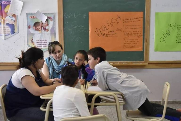 Duas mulheres brancas sentadas à mesa com três crianças brancas em uma sala de aulas