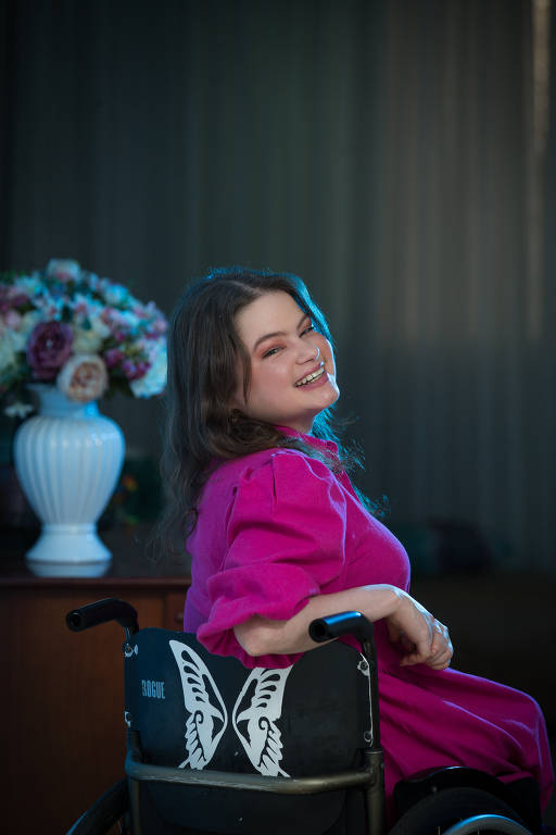 Uma mulher cadeirante, com roupas cor-de-rosa, sorri em um cenário com flores