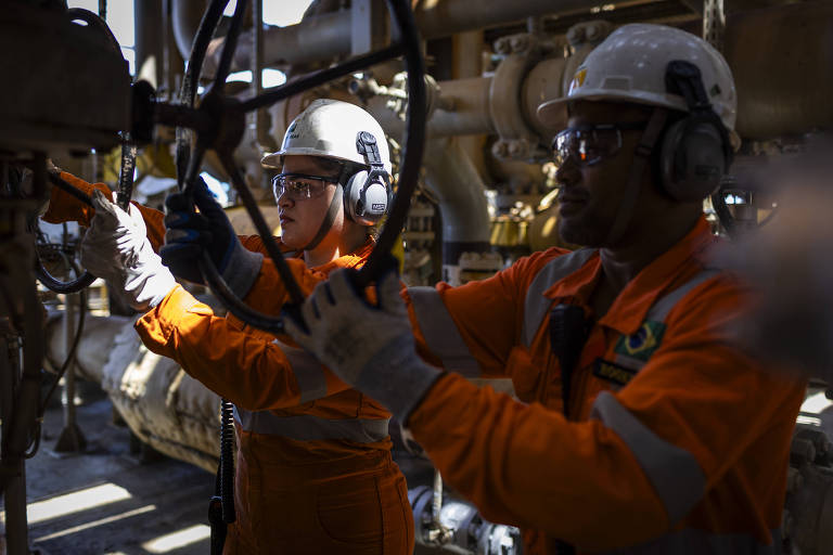 Imagem mostra Dédima, à esq., uma mulher branca que veste equipamento de proteção, um colete laranja, capacete branco com logo da Petrobras e fones cinzas. À direita, seu colega de trabalho veste o mesmo equipamento