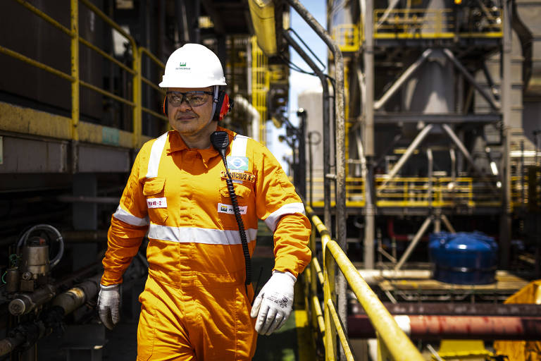 Vinicius é um homem branco que, na imagem, veste equipamento de proteção composto por macacão laranja, luvas brancas, capacete branco com logo da Petrobras e fone laranja. Ele está em meio às construções da plataforma