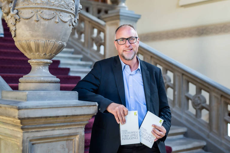 Homem branco de cerca de 40 anos de óculos e terno posa em uma escadaria clássica, segurando dois livros com títulos sobre ética e inteligência artificial; o ambiente sugere uma instituição prestigiosa