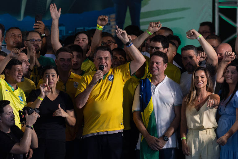 Homem vestido com camisa da seleção brasileira de futebol fala para uma plateia, cercado de apoiadores