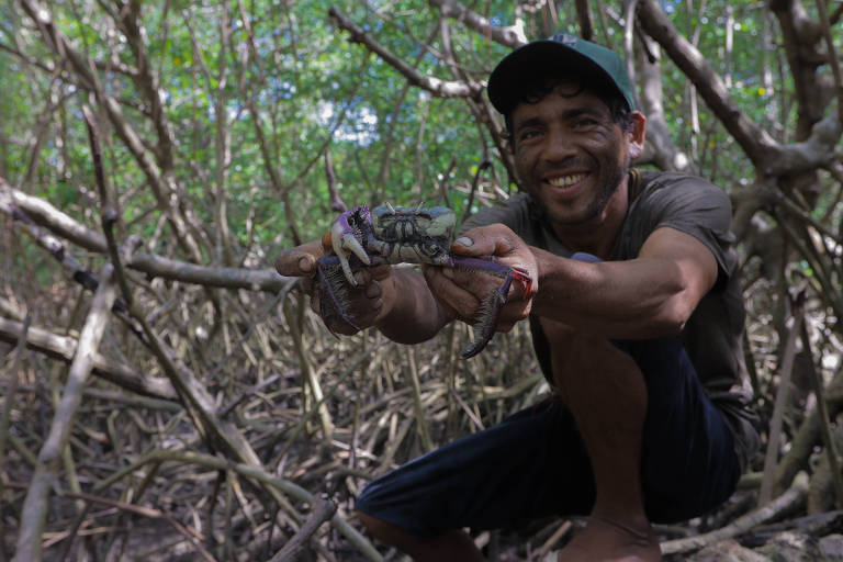 Rogério Matos Souza segura caranguejo com as mãos e sorri em um grande manguezal