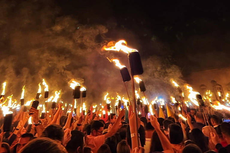 pessoas empunham tochas à noite, iluminando o local com uma luz avermelhada e produzindo névoa de fumaça. a foto é feita por trás da multidão, com o céu escuro ao fundo 