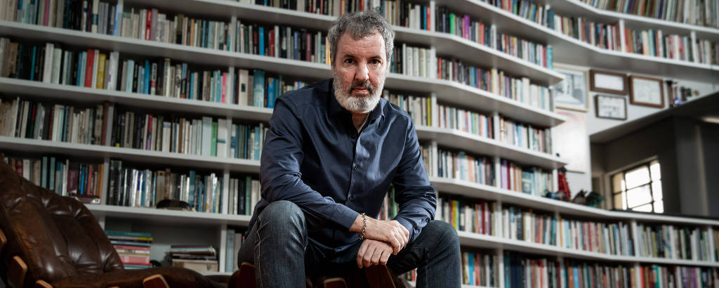 homem branco de cabelo e barba entre preto e grisalho, vestido de azul marinho em frente a estante repleta de livros