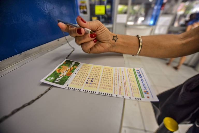 Imagem fechada de um volante da Mega-Sena sobre um balcão e um braço segurando uma caneta para marcar os números