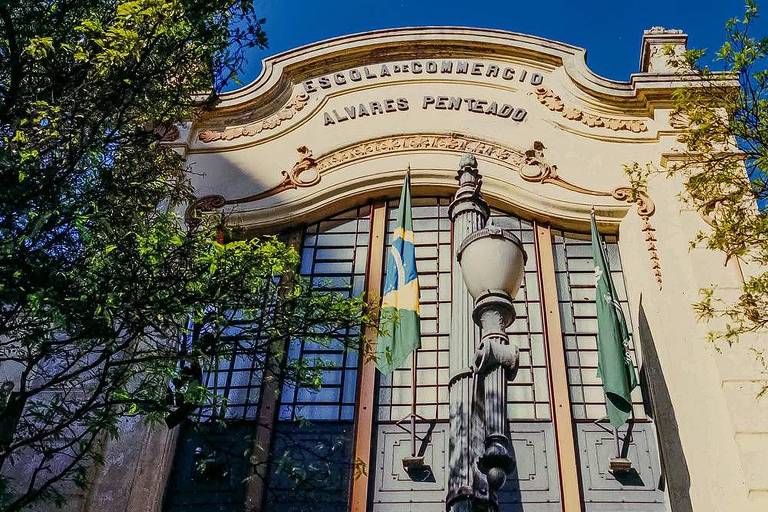 Parede externa de edifício com portas de metal e vidro, com ornamentos e os dizeres "Escola de Commercio, Álvares Penteado", em frente a uma bandeira do Brasil, uma árvore e um poste antigo de luz com desenho antigo