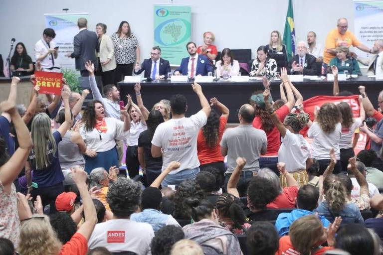 Manifestantes na plateia levantam faixas e erguem as mãos diante de parlamentares sentados no palco na comissão