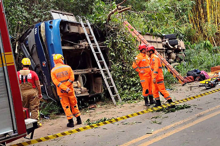 Na foto, quatro pessoas de uniforme laranja estão em uma estrada com uma faixa de interdição, próximos a um ônibus azul tombado em meio à vegetação. Uma escada está apoiada no ônibus, e a parte de trás de um caminhão dos bombeiros aparece à esquerda.