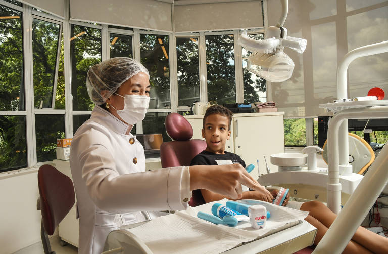 Um menino negro, de 10 anos, está ao lado de uma mulher japonesa, de touca e máscara no rosto; ela veste jaleco branco; a criança olha a mulher mexendo nos aparelhos de um consultório dentário