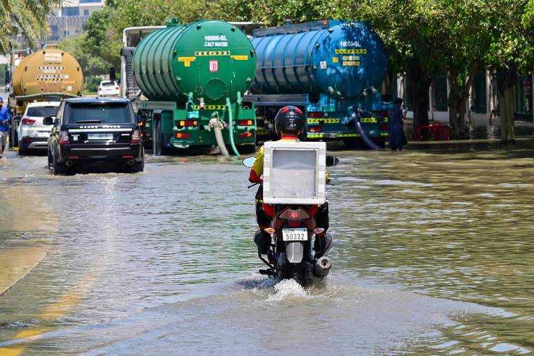 Homem anda de moto em uma rua alagada enquanto três caminhões pipa bombeiam água das inundações 