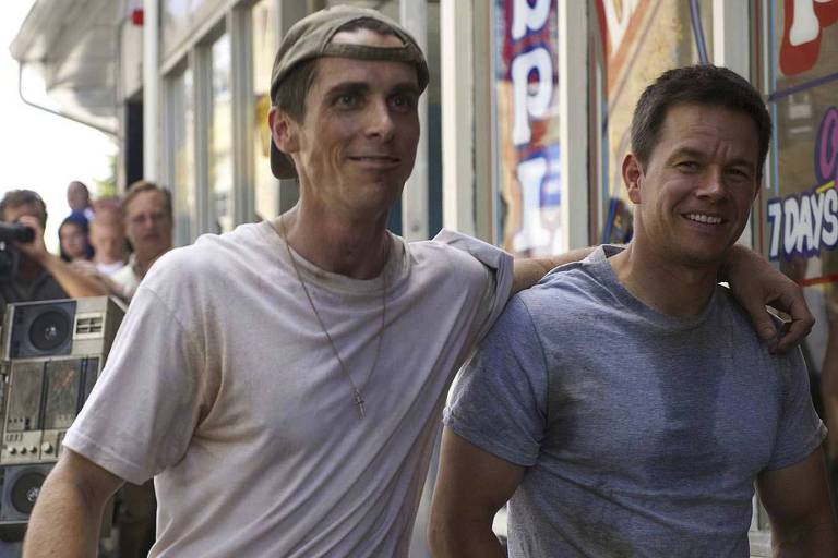 Dois homens sorriem abraçados. O da esquerda, muito magro, veste camiseta branca e um boné virado para trás. O da direita tem a camiseta molhada de suor no peito