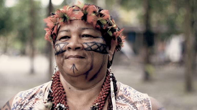 Mulher indígena com pinturas no rosto; ela sorri sem mostrar os dentes, olhando para frente