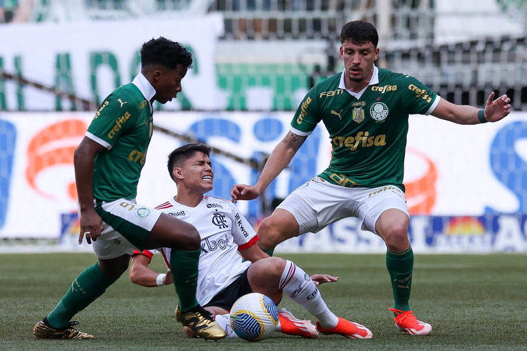 Dois jogadores palmeirenses tentam dominar a bola, enquanto um flamenguista reclama de dor no chão