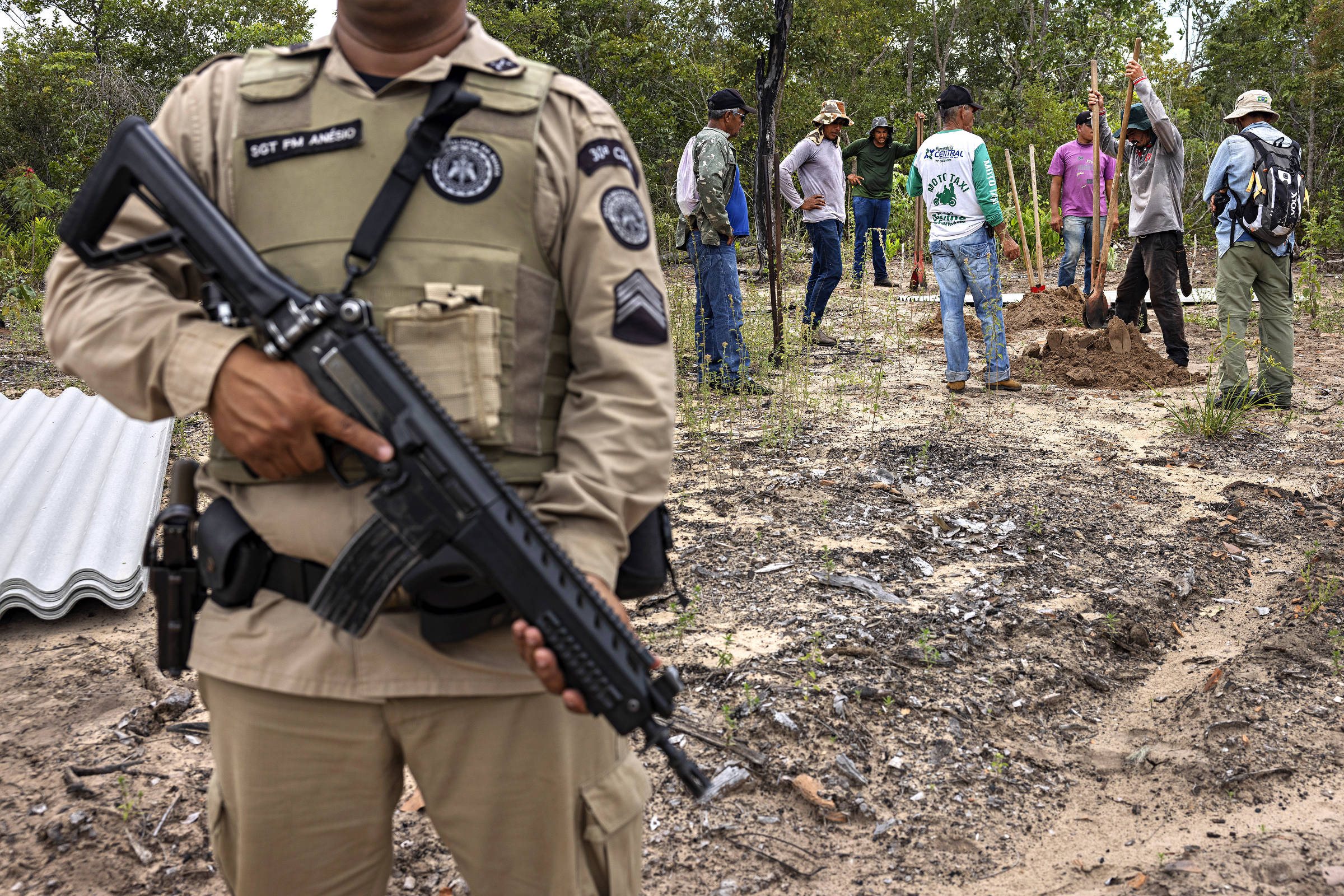 No primeiro plano, policial militar usando colete a prova de balas segura arma; ao fundo, agricultores trabalham na reconstrução de rancho