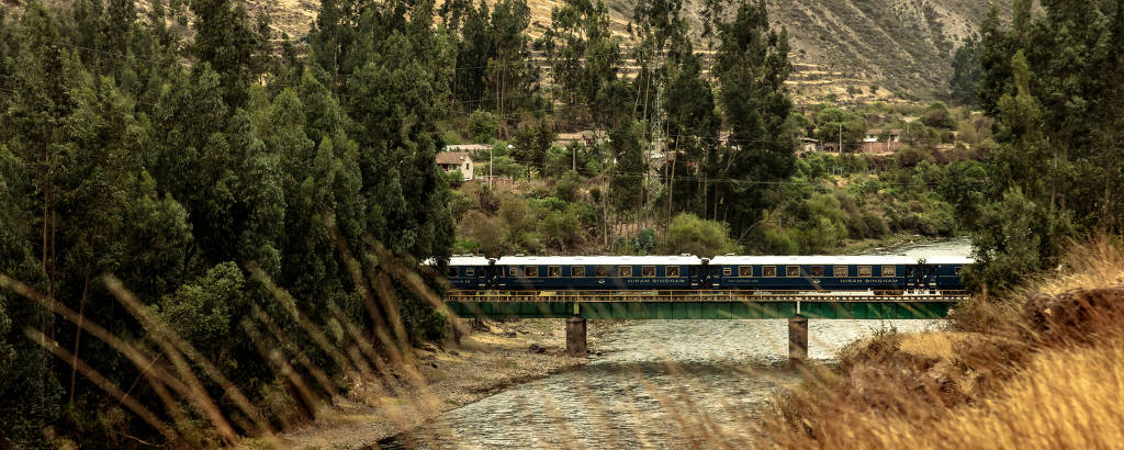 imagem de trem azul passando em ponte em paisagem com árvores e montanhas