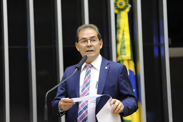 Um homem de terno e gravata está de pé em frente a um microfone, segurando papéis em uma das mãos e gesticulando com a outra. Ele está fazendo um discurso na Câmara, com a bandeira do Brasil ao fundo