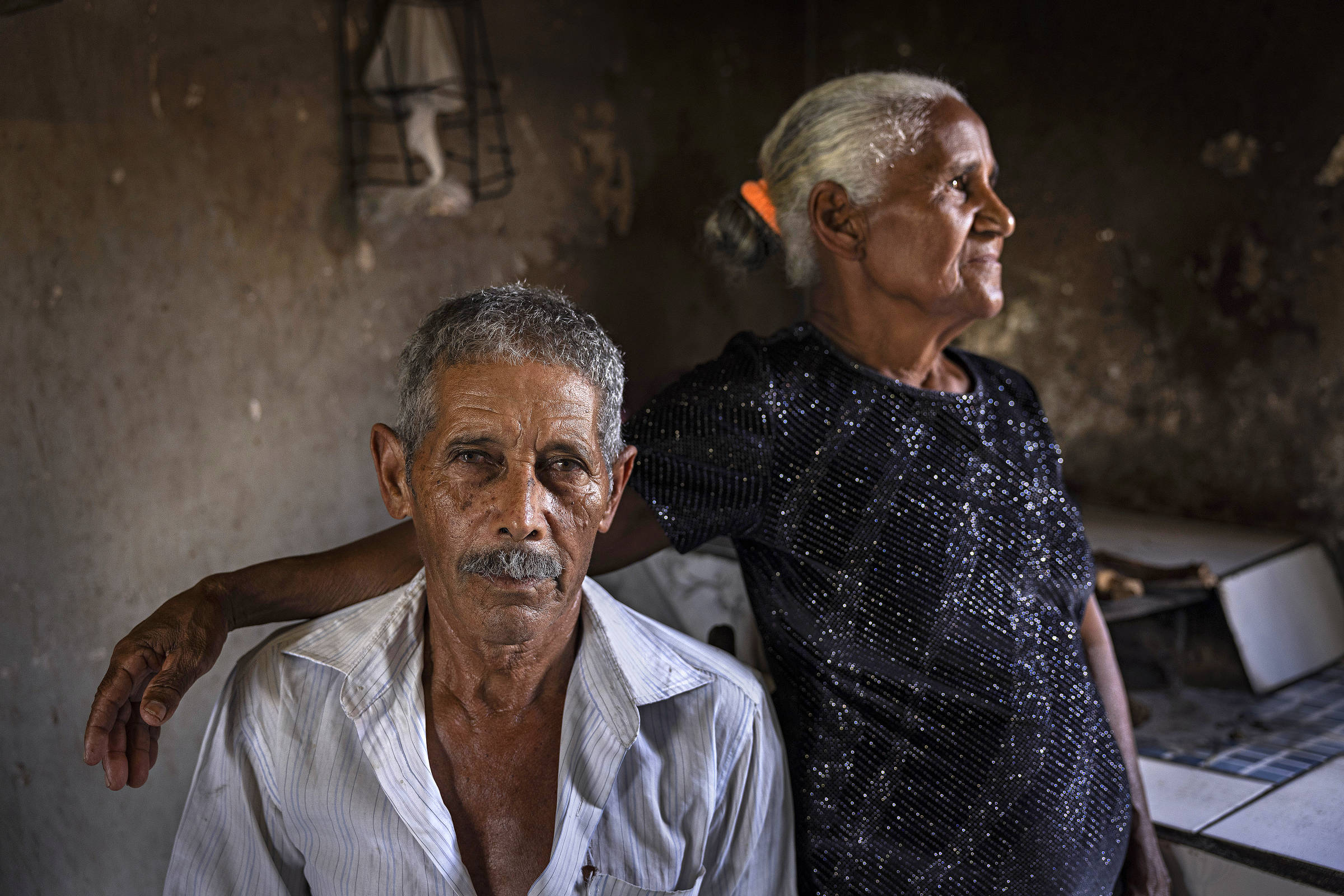 Retrato de homem negro, grisalho, usando camisa branca, sentado ao lado da esposa, uma mulher negra, grisalha, usando vestido azul escuro, que olha para o lado