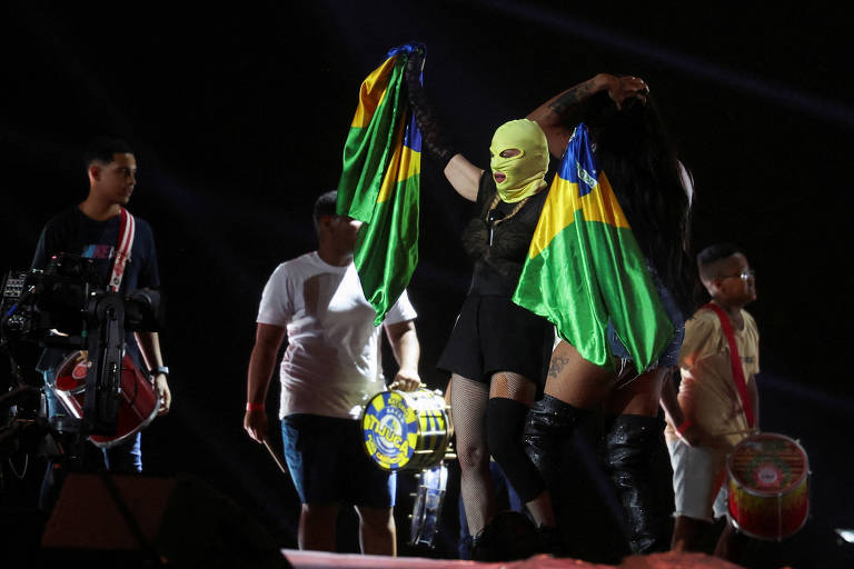 Com máscara verde e roupa preta, a cantora segura duas bandeiras o Brasil no palco