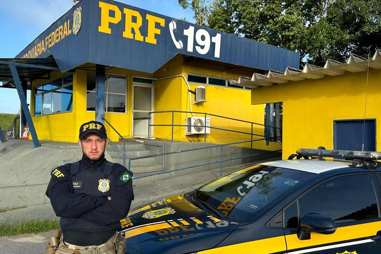 Leandro é um homem branco de barba loira que, na imagem, veste uniforme azul marinho da PRF. Ao fundo, é possível ver uma unidade da polícia e um carro da força