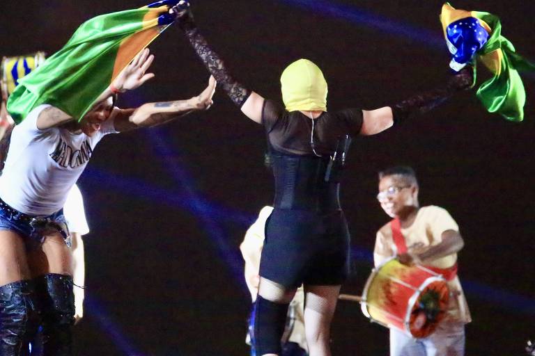 Em foto colorida, mulher dança em cima do palco e ritmistas vibram