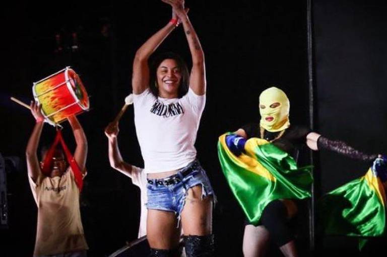 Em foto colorida, mulher dança em cima do palco e ritmistas vibram