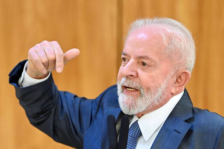 Homem branco idoso vestido de terno azul com camisa branca e gravata azul gesticula com os dedos para baixo enquanto fala; ao fundo, parede com fundo amadeirado mais claro