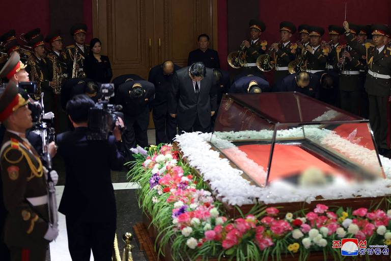 Em destaque, há um caixão rodeado de muitas flores, com o corpo de Kim Ki Nam; ao redor, autoridades e militares prestam homenagens
