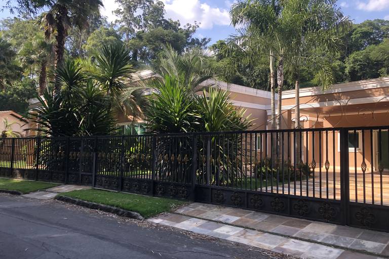 Casa térrea com fachada em tons de bege está cercada por uma cerca de metal preta e um portão. A residência é ladeada por palmeiras e outras árvores. O céu azul com nuvens esparsas sugere um dia ensolarado