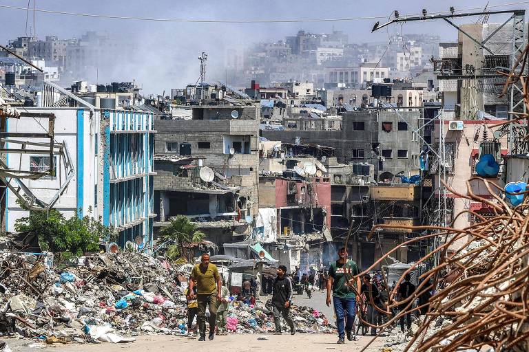 Pessoas passam por monte de lixo e edifícios destruídos enquanto a fumaça sobe durante o bombardeio israelense em Jabalia, no norte da Faixa de Gaza
