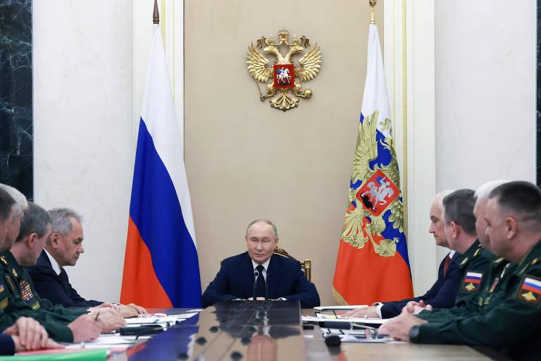 Ladeando Putin em uma reunião do Conselho de Segurança da Rússia estão o ex-ministro da Defesa, Serguei Choigu (esq.) e o novo, Andrei Belousov (dir.), ambos de terno escuro  