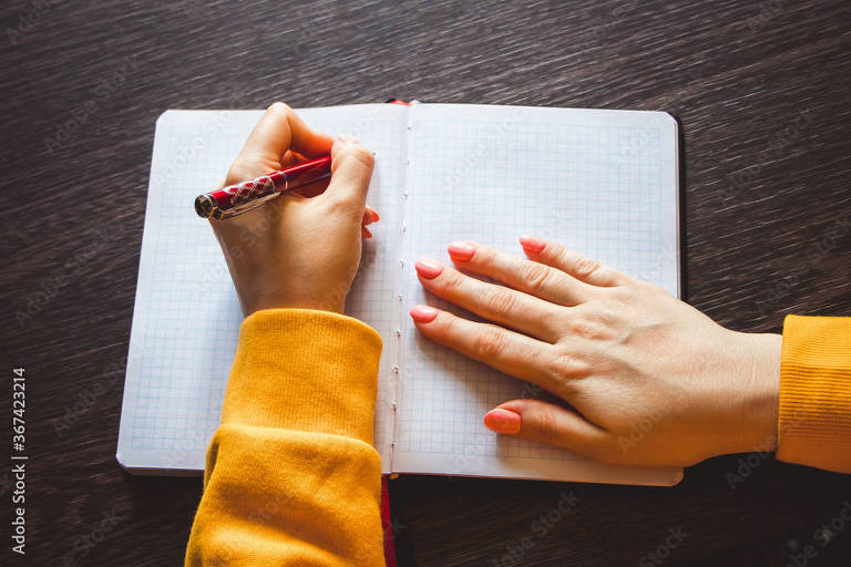 Uma pessoa com as unhas pintadas de vermelho está prestes a escrever em um caderno de capa dura aberto, usando uma caneta vermelha. A pessoa veste uma blusa de manga longa na cor laranja, e o caderno está sobre uma superfície de madeira escura, sugerindo um ambiente de trabalho ou estudo organizado.