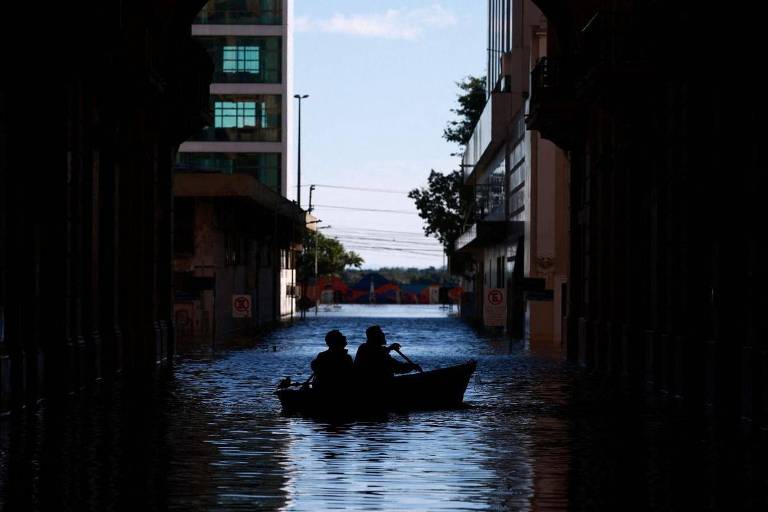 Duas pessoas são vistas navegando em um pequeno barco por uma rua urbana completamente inundada. Os edifícios ao redor estão parcialmente submersos, e a água alcança quase a altura dos sinais de trânsito. A cena é capturada em contraluz, com as silhuetas das pessoas e do barco destacando-se contra o reflexo da água.