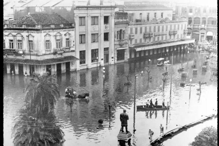Uma rua urbana completamente inundada, com águas refletindo as fachadas dos edifícios históricos. Pessoas navegam em pequenas embarcações improvisadas, enquanto outras observam a cena a partir de passarelas elevadas, evidenciando a gravidade da enchente.