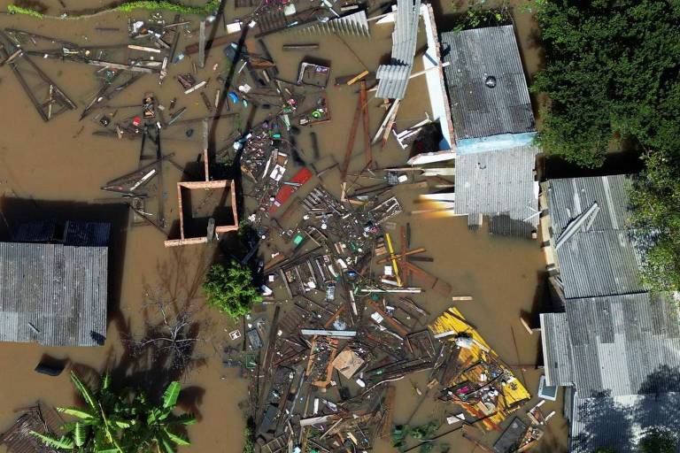 A imagem captura a destruição causada por uma enchente, mostrando casas danificadas e destroços espalhados pela água barrenta. A perspectiva aérea realça o impacto devastador do desastre natural sobre a comunidade.