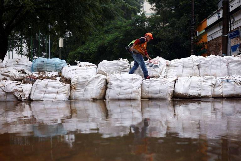 Um trabalhador com capacete e colete refletivo caminha sobre uma barreira de sacos de areia, erguida para conter as águas de uma inundação que cobriu a rua. 