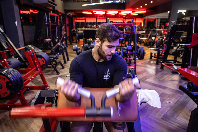 Homem branco sentado em uma máquina de musculação. Ele veste camiseta preta e exercita o bíceps.