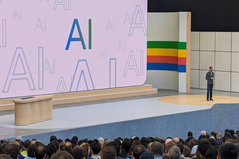 Um orador está em pé em um palco, apresentando para uma audiência em um evento de tecnologia. O fundo do palco exibe a palavra "AI" repetida em vários tamanhos, com um logotipo colorido ao lado, sugerindo o tema de inteligência artificial. Vários espectadores estão na plateia