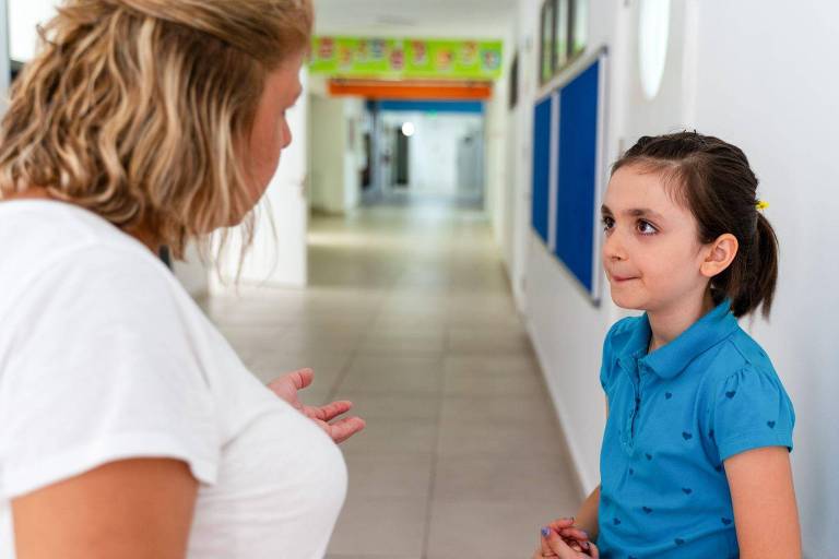 Mulher adulta falando com menina em corredor de escola
