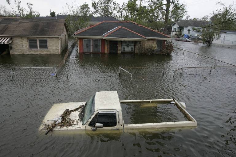 A imagem mostra uma cena de inundação severa em uma área residencial, onde as águas subiram a um nível que submerge parcialmente as casas e veículos. Uma picape branca está quase completamente submerso na água, com apenas a parte superior visível, destacando a gravidade da situação. As casas ao redor também estão cercadas por água.