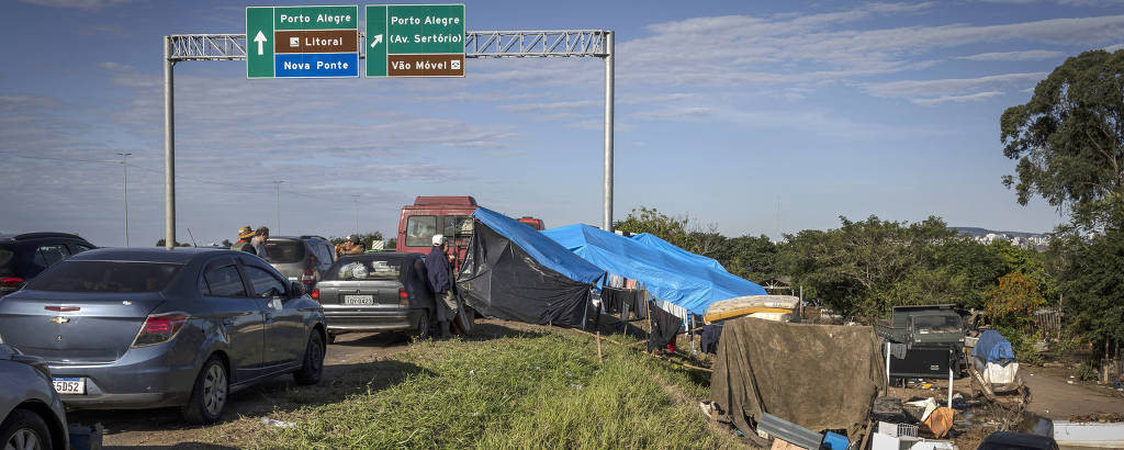 A imagem mostra uma rodovia, onde veículos passam por uma área improvisada com barracas e pertences espalhados, indicando uma situação de moradia precária. Um grande letreiro de trânsito ao fundo indica direções para cidades