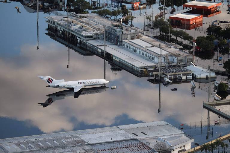 Na foto, um avião está estacionado em uma pista alagada, que reflete as nuvens no céu. Próximo ao avião está o aeroporto, também alagado.