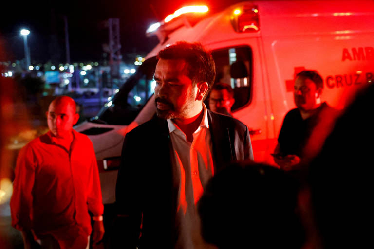 Um homem de expressão séria caminha à noite, iluminado por luzes vermelhas e amarelas, com uma ambulância da Cruz Vermelha ao fundo, sugerindo uma cena de emergência médica.
