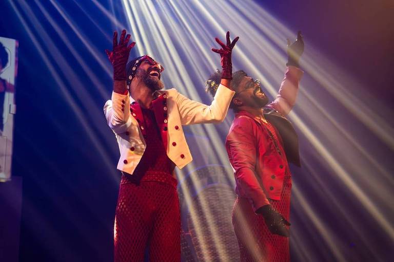 Dois artistas vestidos com trajes vermelhos e brancos, apresentam-se sob holofotes em um palco