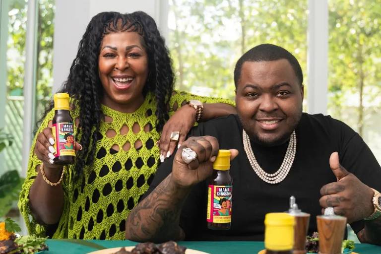 O cantor Sean Kingston, um homem negro de cabelo raspado, segura um vidro de molho e faz sinal de joinha com a mão ao lado e sua mãe, Janice Turner, à esquerda, uma mulher negra de cabelos cacheados que veste roupa amarela